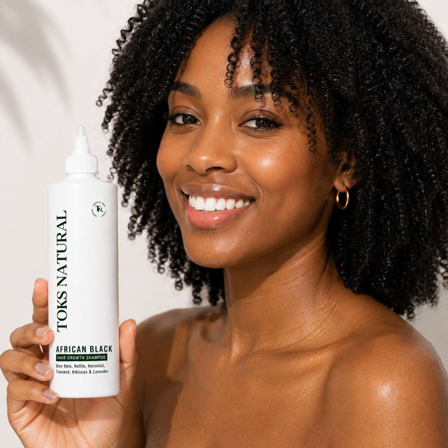 A smiling woman with dark curly hair holds up a white bottle of TKS Natural African Black hair developer against a clean white background.