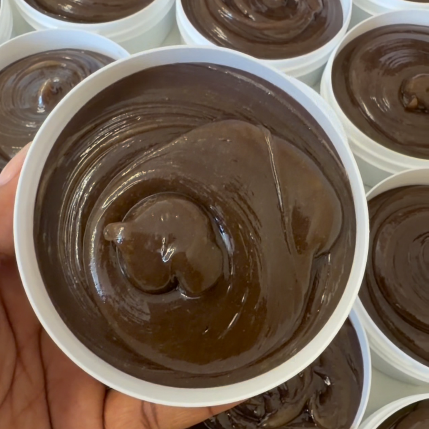 A close-up of a hand holding a white cup filled with thick, glossy dark brown chocolate spread, with multiple similar cups arranged in the background.