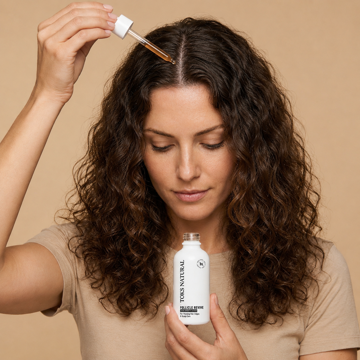 A woman with long, curly brown hair holds a dropper bottle of Toks Natural Hair Growth Serum, applying serum from the glass dropper to her hairline. She is wearing a beige t-shirt against a solid beige background.