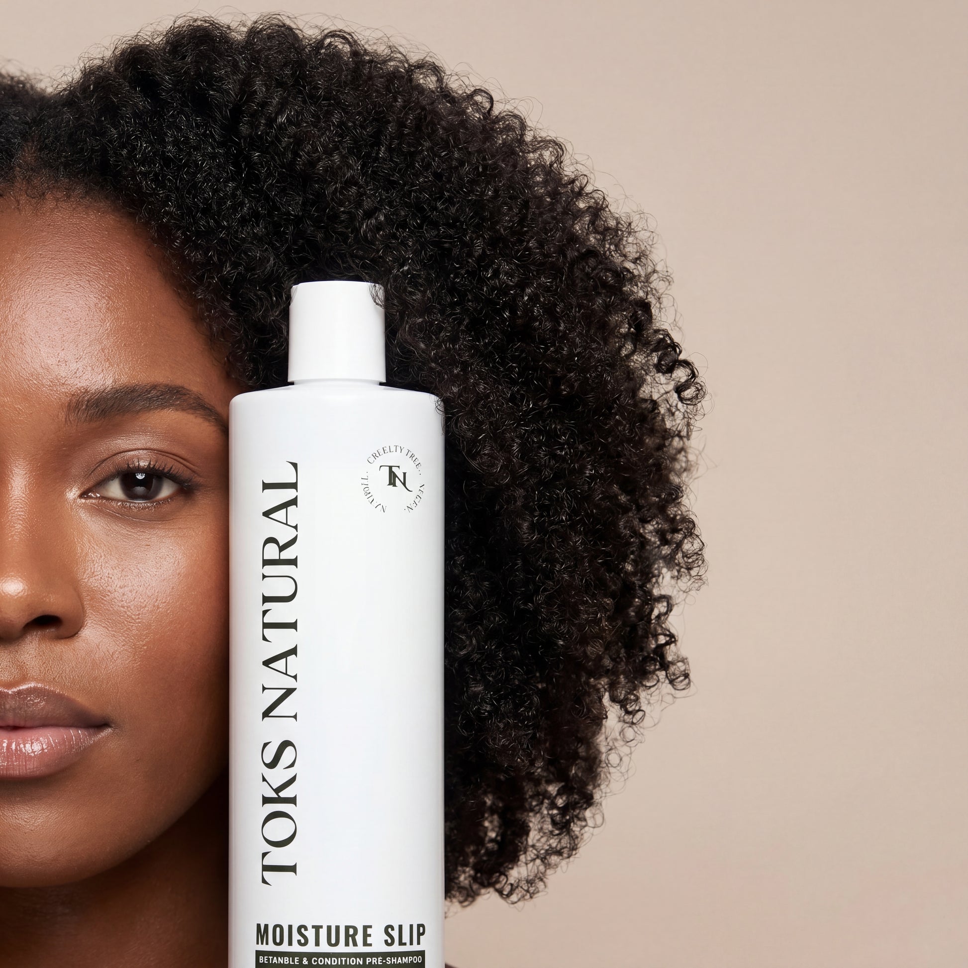 Woman with curly hair holding a bottle of TOKS Natural hair product against a beige background