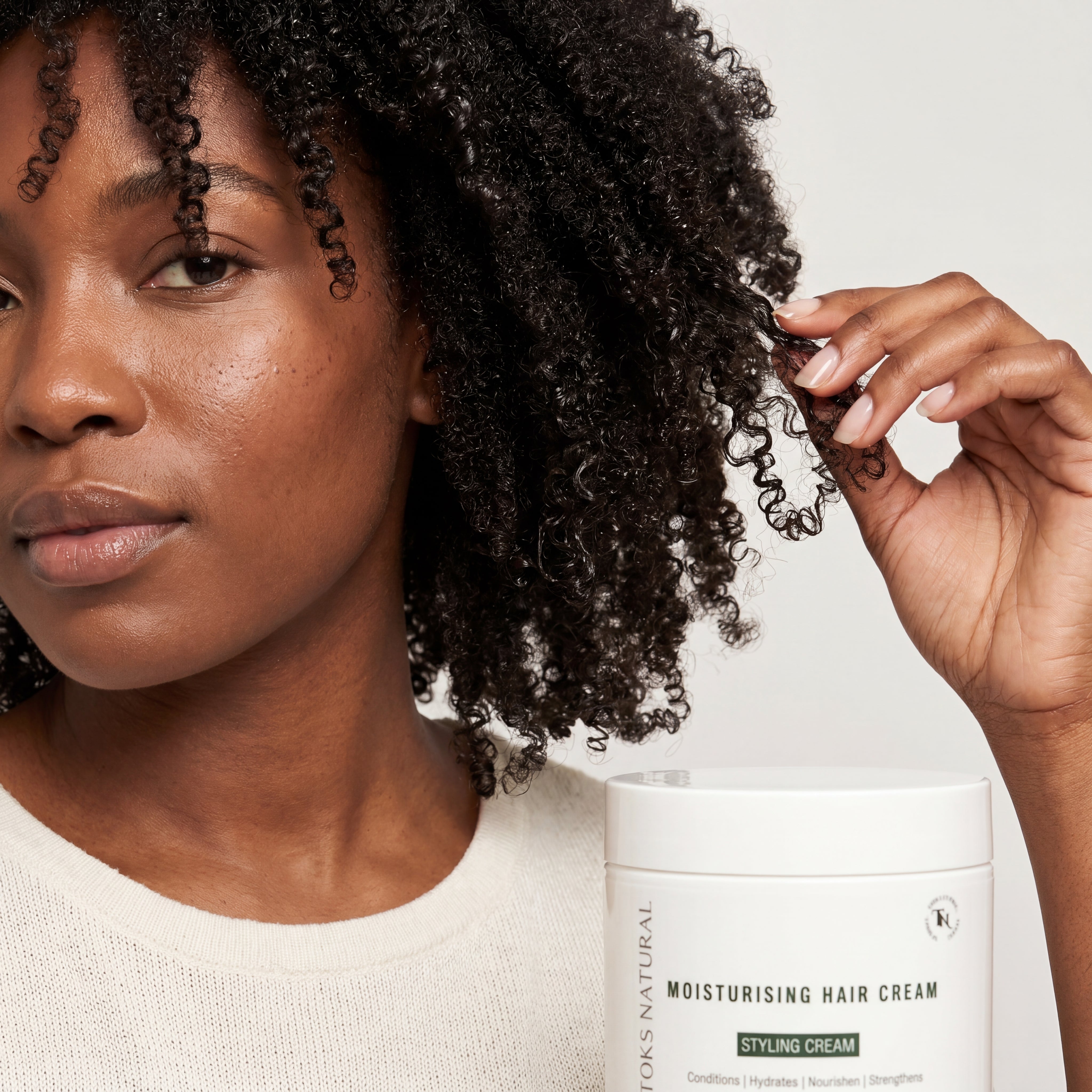 Woman with curly hair touching a container of Toks Natural Styling Cream on a neutral background