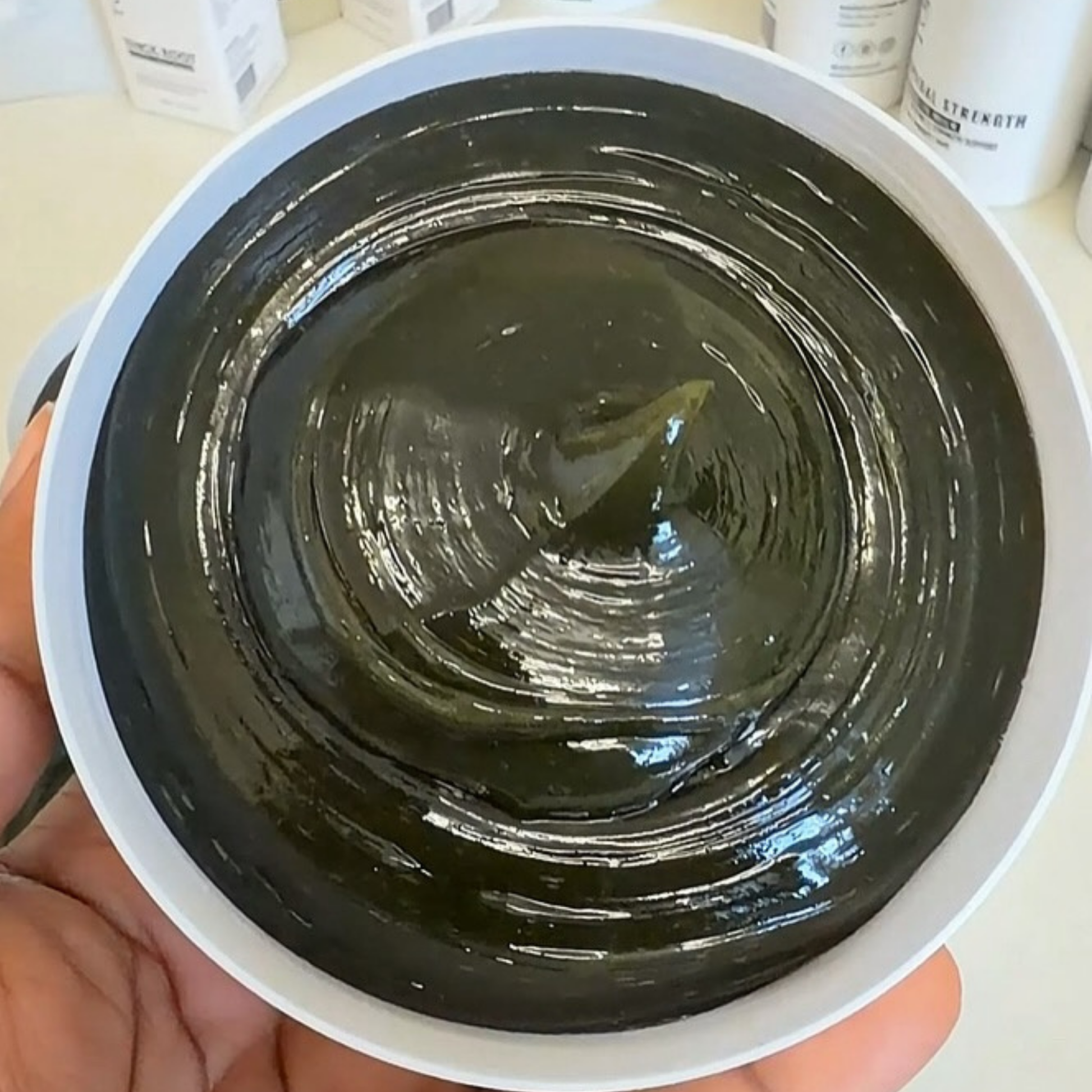A close-up of a hand holding a white bowl filled with a thick, glossy black cosmetic or skincare paste, with blurred product containers in the background.